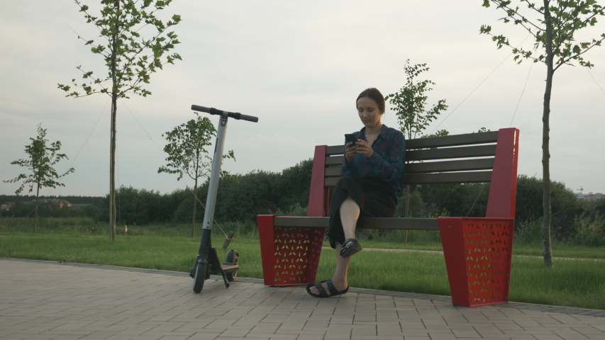 Happy successful businesswoman sits on bench with electric scooter in park. Businesswoman with alternative electric transport. Young beautiful woman with is sitting in park with phone in her hands