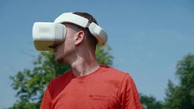 Man in virtual reality helmet is studying virtual game. Guy in the park in red T-shirt on background of blue sky and green trees looks VR glasses. 4K footage