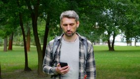 Gray-haired, focused man travel blogger records video sightseeing on smartphone for social media subscribers. Guy tourist in shirt, backpack walks in park makes photos on smart phone. Slow motion