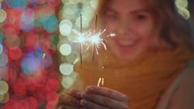 Blonde woman smiling holds Christmas lights sparklers splashes burn in hands Christmas song against of bokeh of Christmas lights in city square in knitted warm orange scarf winter. Noel. Emotions