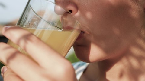 Woman is drinking orange juice. Side view of female hand bring glass with orange juice to their mouth and drink it on sunny day in nature. Close-up