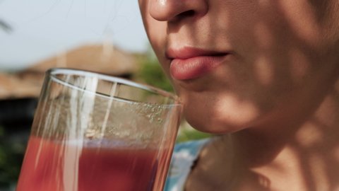 Woman is drinking tomato juice. Side view female hand bring glass with tomato juice to their mouth and drink it on sunny day in nature. Close-up