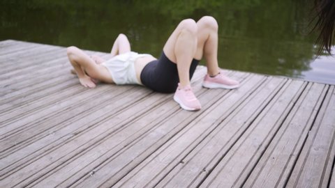 slim young sportswomen in tracksuits do exercises bending over backwards and rest on wooden pier near pound water in city park on summer day closeup