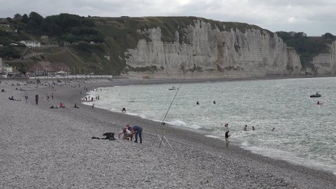 FECAMP, FRANCE – JULY 2020: 	People swim and fish at rocky beach of Fecamp, with beautiful white cliffs in the background, holiday destination in France
