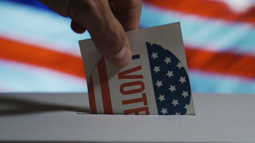 USA elections 2020, the hand of a man putting his vote in a ballot box with a waving US National flag on the background Royalty-Free Stock Footage #1057449979