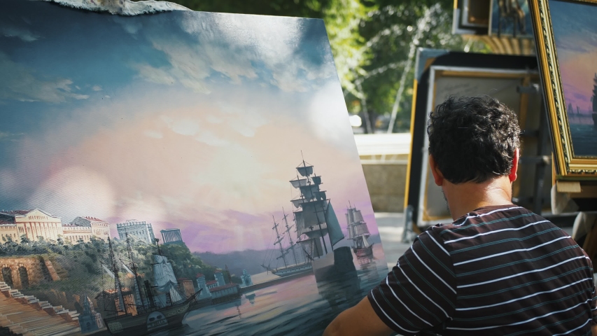 Male artist drawing a picture with marine landscape, sailing ships and city on coast while sitting in park