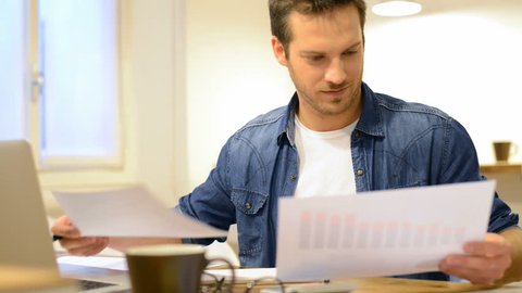 Happy young businessman reading paperwork at desk in office. Casual man smiling at work. Young man working at business document and checking the file on his laptop. Man working at documents.