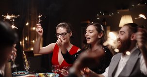 Group of multiethnic friends celebrating christmas together, cheering, smiling and holding burning sparklers at dinner party table - celebration, real people concept 4k footage