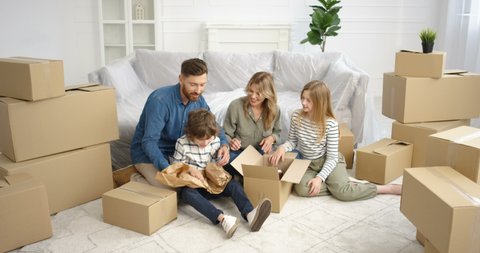 Cheerful Caucasian parents sitting on floor in living room with small cute kids and unpacking carton boxes. Family with children moving in new house after repairment Boy and girl with mother ad father
