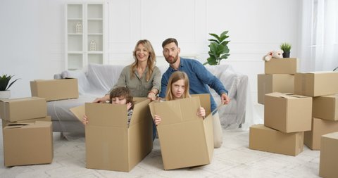 Happy Caucasian parents having fun in living room with kids while moving in new house. Cute little children, boy and girl sitting in carton boxes and playing with mother and father in new apartment.