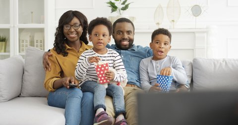 Happy cheerful African American family sitting on couch in living room and watching TV together with popcorn. Cute kids with smiled joyful mother and father having fun time together at home. Parenting