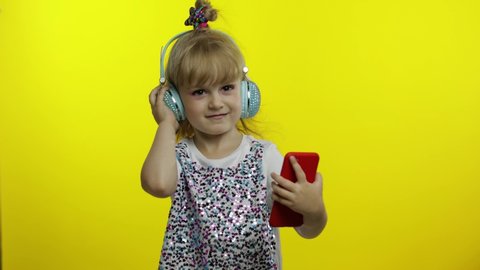 Child dancing with smartphone, listening to music on headphones. Energetically moving to the rhythm, relaxing, enjoying, having hun. Little fun blonde kid girl isolated on yellow background in studio