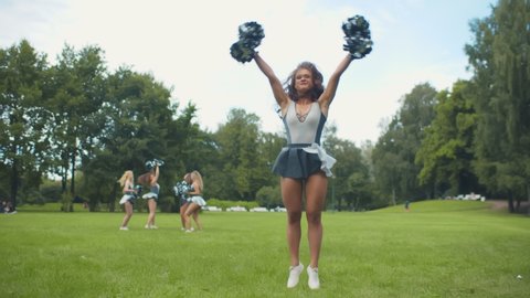 Portrait of cheerleader in uniform jumping in air with pompons practicing before performance. Active cheerleading girl doing mid-air split with teammates training on background