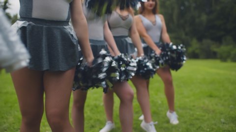 Cropped shot of cheerleading team in uniform performing dancing element with pompons preparing for competition and training outdoors in park.