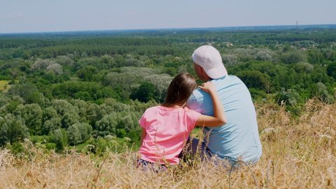beautiful summer landscape. man and little girl, dad and daughter, sit on cliff edge, admiring amazing panorama of green valley, and blue sky with rare clouds. Back view.