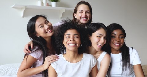 Head shot attractive young mixed race diverse girls sitting on bed, looking at camera. Happy smiling millennial female friends students enjoying pajama sleepover slumber party together in bedroom.