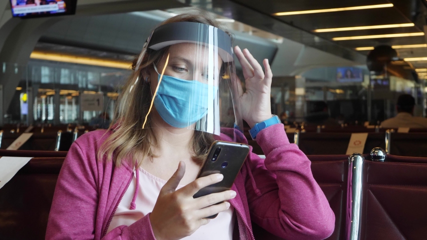 Woman in blue medical mask and protective face shield in airport. Female use Internet on smartphone during wait flight. Prevent coronavirus COVID-19 infection. Protection measures during pandemic
