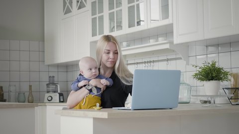 Mother multi-tasking, holding baby infant and using computer laptop at home. Adorable young woman kisses her toddler son while working with computer. Young Woman Taking Care Of Her Baby While Working