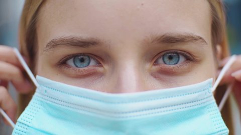 Close up face young woman with blue eyes stand looking at the camera wearing medical mask being outdoors during quarantine pandemic covid-19 slow motion