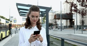 Front view of confident young lady in eyewear and white shirt walking on street and using modern smartphone. Beautiful girl with wavy brown hair browsing internet outdoors.
