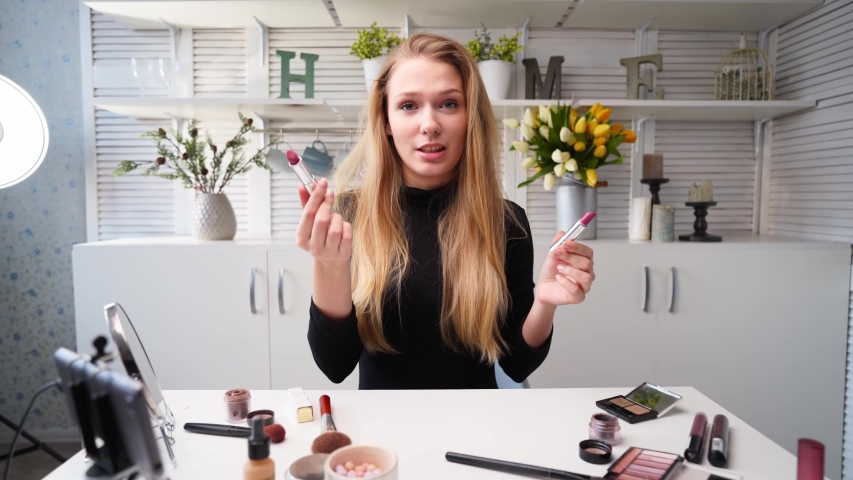 Vlogger female applies lipstick on lips. Beauty blogger woman filming daily makeup routine tutorial at camera on tripod. Influencer blonde girl live streaming cosmetics product comparison in studio | Shutterstock HD Video #1057479847