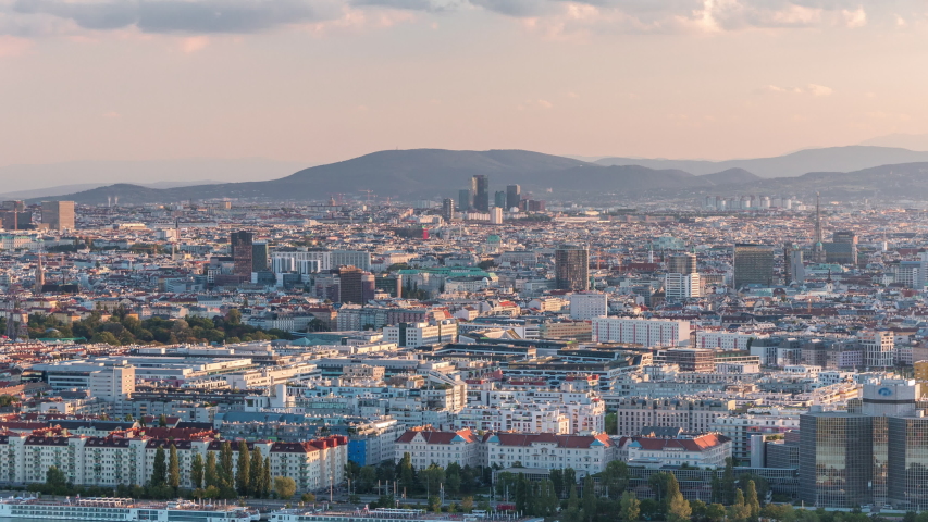 Aerial panoramic view of Vienna city with skyscrapers, historic buildings and a riverside promenade timelapse in Austria. Evening skyline before sunset from Danube Tower viewpoint. Mountains on a