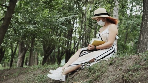 The young woman with a hat on her head and a medical mask on her face sits on the ground in the park while taking notes in her notebook and looking at the lake in front.