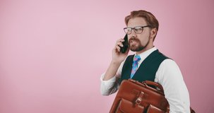 Successful businessman in eyeglasses and suit talking on smartphone over pink background. Mature man holding brown briefcase during conversation in studio.