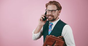 Cheerful man wearing eyeglasses and formal clothing, having phone conversation over pink background. Bearded businessman with leather briefcase standing in studio.