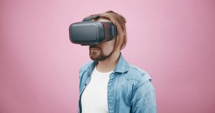 Handsome man in denim shirt experiencing virtual reality in VR headset. Isolated over pink studio background. Concept of people and modern technology.