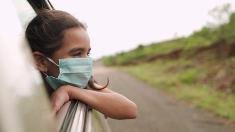 Sad Little girl kid with medical mask looking out from moving car window during coronavirus or covid-19 pandemic - Concept of new normal, lifestyle and travel.