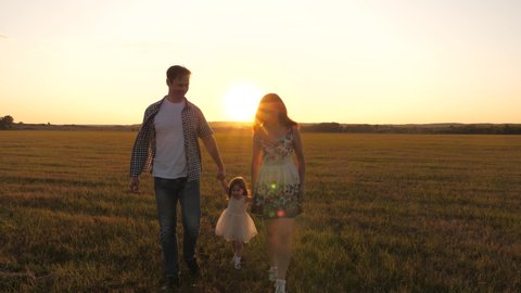 little daughter holds mom and dad by hands and jumps on meadow in park in rays of yellow sun. Family happiness. healthy baby playing with dad and mom on field in sunset light. Walking child in nature.