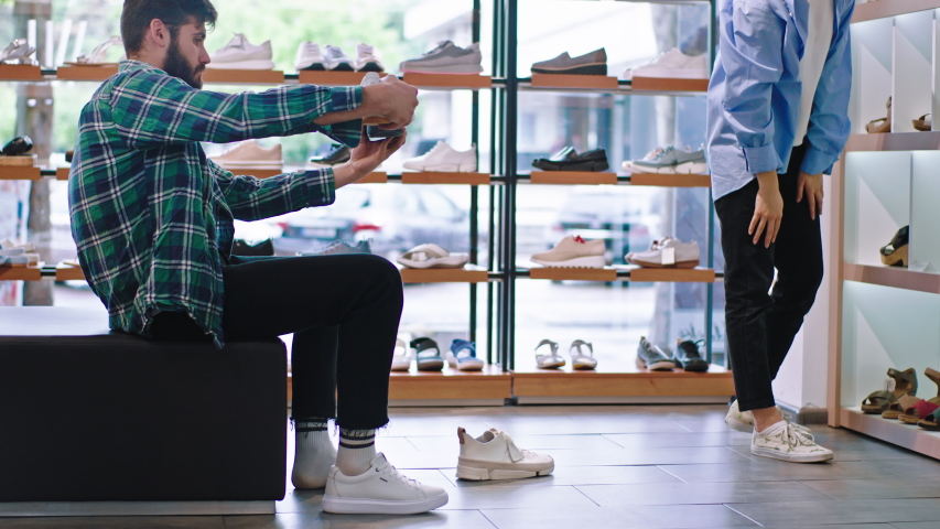 Good looking customer guy take a sit on the sofa in the shoe shop he make difference between shoes he wants to buy.  Royalty-Free Stock Footage #1057495192