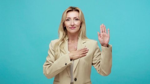 Responsible adult blond woman in business style suit holding hand on chest swearing and promising, proud to be part of her country, patriotism. Indoor studio shot isolated on blue background