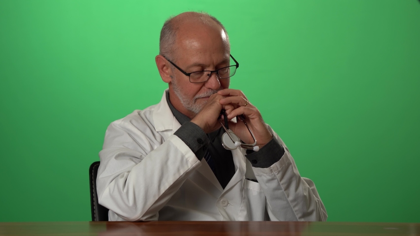 Male doctor wearing a lab coat and stethoscope on green screen sitting at desk dealing with depression or grief. Royalty-Free Stock Footage #1057497124
