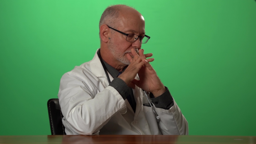 Male doctor wearing a lab coat and stethoscope on green screen sitting at desk dealing with depression or grief. Royalty-Free Stock Footage #1057497193