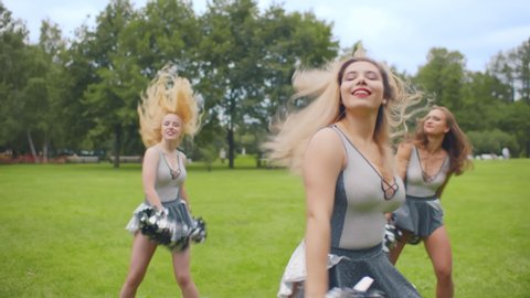Attractive young cheerleaders working out in summer park. Group of group of cheerleading girls in white and grey uniform dancing with pom-poms in their hands outdoors