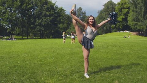 Beautiful cheerleader in uniform practicing split holding pompom standing outdoors. Portrait of cheerleading girl stretching and warming up training in park