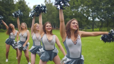 Portrait of beautiful cheerleader girls in white-grey uniform standing in line and dancing with pompoms outdoors. Cheerleading team practicing outdoors