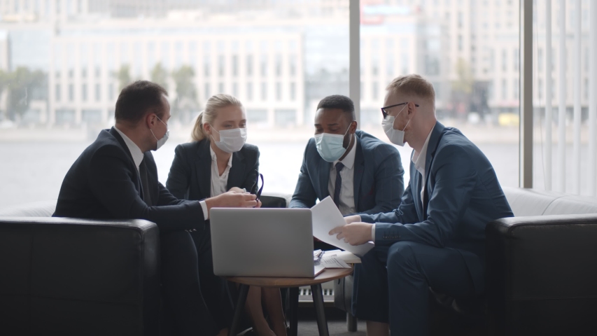 Diverse business team in protective masks working in modern office. Multiethnic colleagues wearing medical masks using laptop discussing new financial strategy sitting on couch Royalty-Free Stock Footage #1057501081