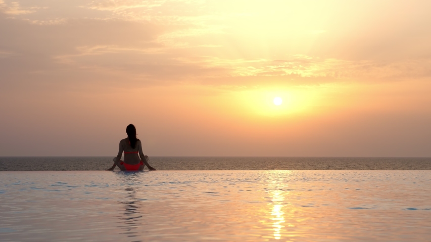Back view, silhouette of woman in swimsuit, doing yoga, meditating, relaxing with hands up, on edge of outdoor infinity pool with panoramic sea view, at sunrise. | Shutterstock HD Video #1057501999