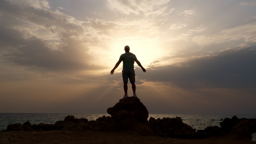 Dark male silhouette, man stands on rocks in front of the ocean or sea, with his arms outstretched and hands up at dawn, sunrise. concept of human strength and faith | Shutterstock HD Video #1057502086