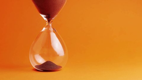 Shot of a sand clock measuring time while the sand is falling down against the orange background - old classic timer. Extreme close up of a transparent hourglass with flowing black sand - time concept