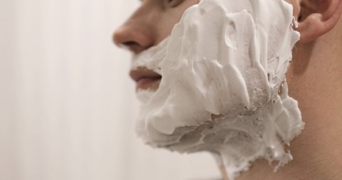 Young man is shaving on cheek with foam using disposable razor in bathroom, closeup, side view. Daily morning routine, hygienic procedure. Man is shaving beard and moustache, part of face in frame.