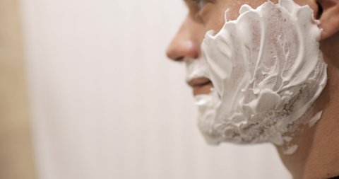 Young man is shaving using disposable razor in bathroom, closeup, side view. Daily morning routine, hygienic procedure. Man is shaving beard and moustache, part of face in frame.