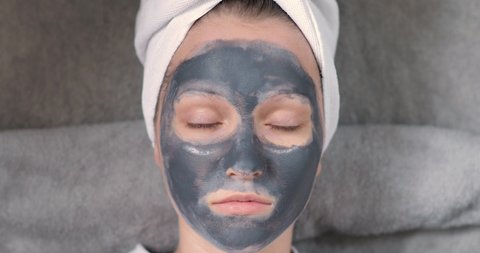 Young woman with grey mask on face is putting slices of cucumber on eyes. Her hairs wrapping in white towel. Spa beauty procedures at home for girl. Skincare, hydrating and moisturising concept.