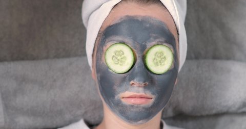 Young woman with clay mask on face taking cucumber from eye and eating it during spa procedure at home. Her hairs wrapping in white towel. Skincare, hydrating and moisturising concept. Joke, humor.