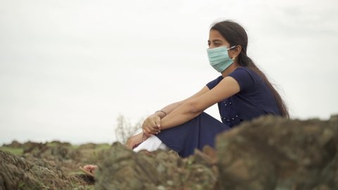 Sad young girl in medical mask sitting on top of mountain nature after coronavirus pandemic - concept of lonely, adhd, lost hope during covid-19