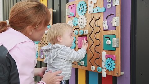 Mother And Child By Busy board. Cute baby boy playing with busy board on the wall. Educational toys. Busy-board for children. Wooden game board