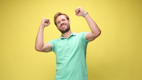 enthusiastic casual man looking up, holding fists in the air and cheering victory, laughing and screaming on yellow background
